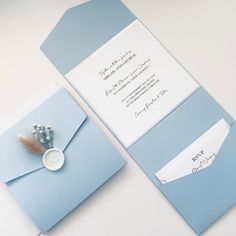 a blue and white wedding card with a button on the front is laying next to an envelope