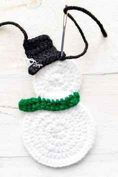 a crocheted snowman ornament hanging on a white wooden background with green trim