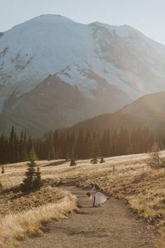 a bride and groom walking down a dirt path in front of a snowy mountain range