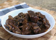 a white bowl filled with meat and mushrooms on top of a wooden table next to a napkin