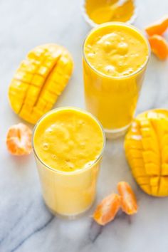 three glasses filled with orange juice on top of a marble counter next to sliced mangoes