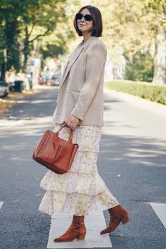 a woman is walking across the street carrying a brown purse and wearing a tan cardigan