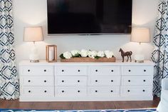 a television mounted on the wall above a white dresser with flowers and a horse figurine