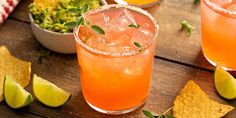 two cocktails with garnish, lime and chips on a table next to a bowl of guacamole