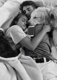 a man and woman laying in bed next to a dog with an electronic device on their lap