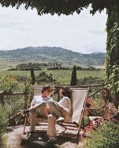 two people sitting on a bench in the middle of an outdoor area with flowers and greenery