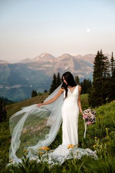 a woman in a wedding dress standing on top of a grass covered hill with her veil blowing in the wind