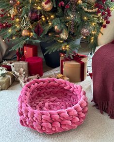 a christmas tree with presents under it next to a pink knitted basket and wrapped gift boxes