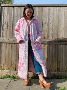 a woman standing in front of a wooden fence wearing a pink and white coat with flowers on it