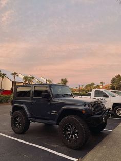 a black jeep parked in a parking lot next to other vehicles and palm trees at sunset