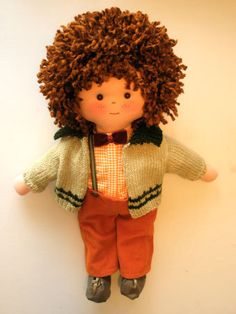 a small doll with curly hair wearing an orange pants and jacket, standing against a white wall