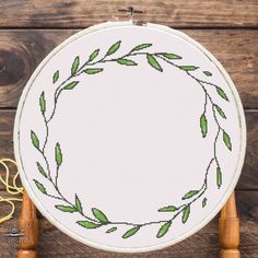 a cross stitch wreath with green leaves in the center on a wooden table next to scissors and thread