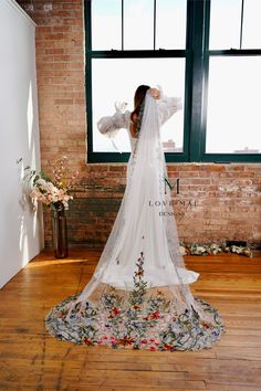 a woman standing in front of a window wearing a wedding dress with flowers on it