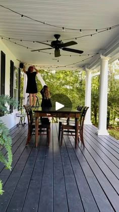 a woman standing on top of a wooden porch next to a table and chair under a ceiling fan