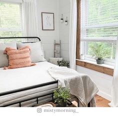 a bed with white sheets and pillows in a bedroom next to two windows that have plants on them