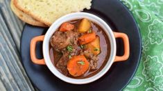 a bowl of beef stew with carrots and bread on a black plate next to a green tablecloth