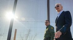 two men in suits and sunglasses walking behind a fenced off area with barbed wire