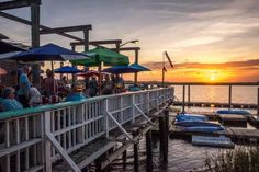 people are sitting at tables near the water as the sun sets over the ocean behind them