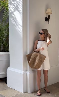 a woman leaning against a pillar holding a straw bag and wearing sunglasses, with palm trees in the background