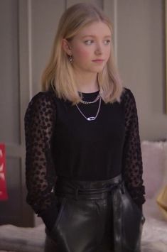 a woman standing in front of a bed wearing a black top and leather skirt with sheer sleeves