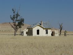 an old abandoned house sits in the middle of a dry grass field with trees and hills in the background