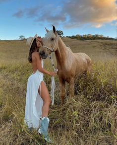 a woman in a white dress standing next to a brown horse on a dry grass field