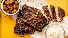 a yellow tray topped with ribs and rice next to two bowls of food on top of it