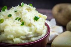 mashed potatoes in a red bowl with green onions on the side and sliced potatoes next to it