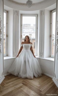 a woman in a white wedding dress standing by a window with her hands on her hips