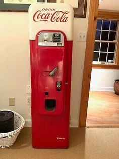 an old fashioned coca cola machine sitting on the floor in front of a wall with pictures above it