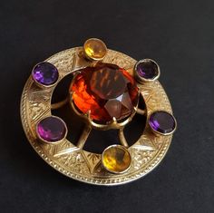 A large, beautiful, and richly saturated antique Scottish Cairngorm paste citrine, tartan brooch. Circa 1870s this Victorian plaid sash pin features a 9k hand engraved gold base, with a halo of amethyst, citrine, and garnet pastes orbiting a vibrant cognac colored center referencing regional stones mined from the Cairngorm mountains.  Brilliant and round the center faceted stone is held by prongs in a crown like setting where it showcases a warm gleaming fire. The surrounding flat cut stones ris Tartan Brooch, Scottish Jewelry, Tartan Sash, Scottish Jewellery, Special Necklace, Victorian Gold, Lovely Shop, Victorian Jewelry, Antique Jewellery