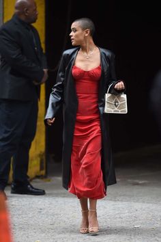 a woman in a red dress and black coat is walking down the street with a handbag