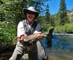 a man holding a fish while standing on a river bank in front of another person