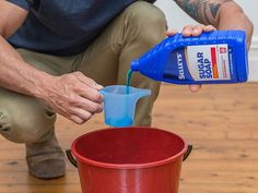 a man pouring water into a red bucket on top of a wooden floor next to a blue container