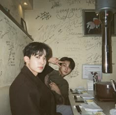 two people sitting at a table with writing on the wall behind them and one person brushing his hair