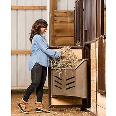 a woman standing next to a horse holding hay in it's back pocket and looking into the stall