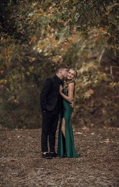 a man and woman standing next to each other in front of trees with leaves on the ground