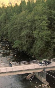 a car driving across a bridge over a river next to a lush green forest covered hillside