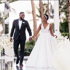 a man and woman walking down the aisle holding hands in front of an archway with white flowers