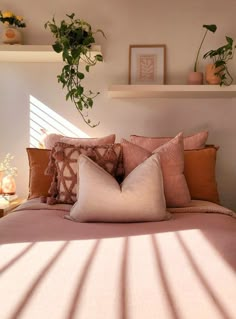 a bed with lots of pillows and plants on the headboard, in front of a window