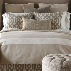 a bed with beige and white comforter, pillows and footstools in front of a brown headboard