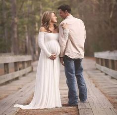 a pregnant couple standing on a bridge in the woods with their arms around each other