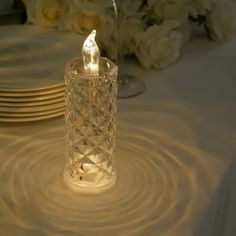 a clear glass candle holder sitting on top of a white table cloth with flowers in the background