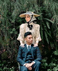 a man sitting next to a woman in a dress and hat on top of her head