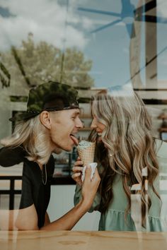 a man and woman sitting at a table eating ice cream cones in front of a window