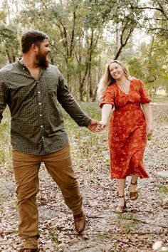 a man and woman holding hands walking through the woods with leaves on the ground in front of them