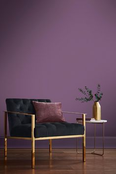 a chair and table in front of a purple wall with a gold vase on it