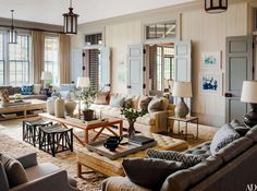 a living room filled with lots of furniture next to two large windows and lamps hanging from the ceiling