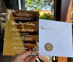 a person holding up a wedding card in front of an open envelope with a gold seal on it