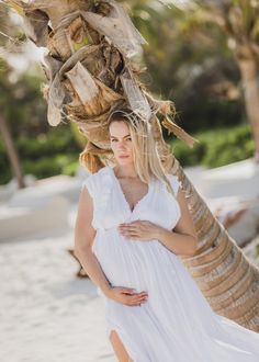 a pregnant woman in a white dress standing on the beach with palm trees behind her